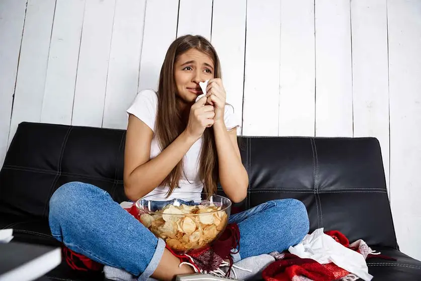 Sad young beautiful girl watching melodrama, crying, sitting on sofa at home. Copy space.