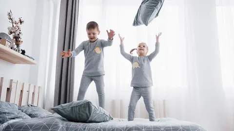 Happy kids playing in white bedroom. Little boy and girl, brother and sister play on the bed wearing pajamas. Nightwear and bedding for baby and toddler. Family at home.